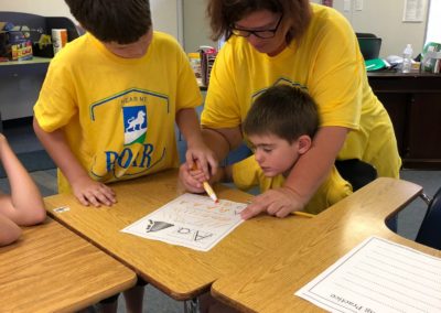 Teacher helping a boy learn to write while another boy watches